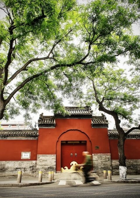 An elderly man is riding his bicycle past a traditional red Chinese gate.