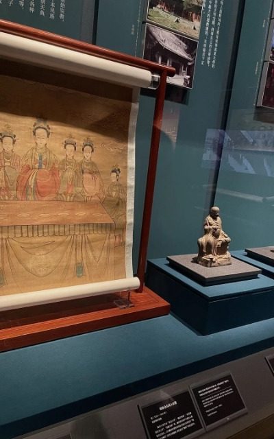 The museum display case features an ancient Chinese painting and two small Buddha statues.